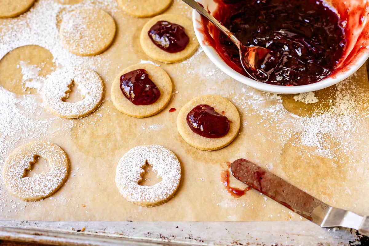 unassembled cookies with jam and powdered sugar on a tray with a bowl of jam.