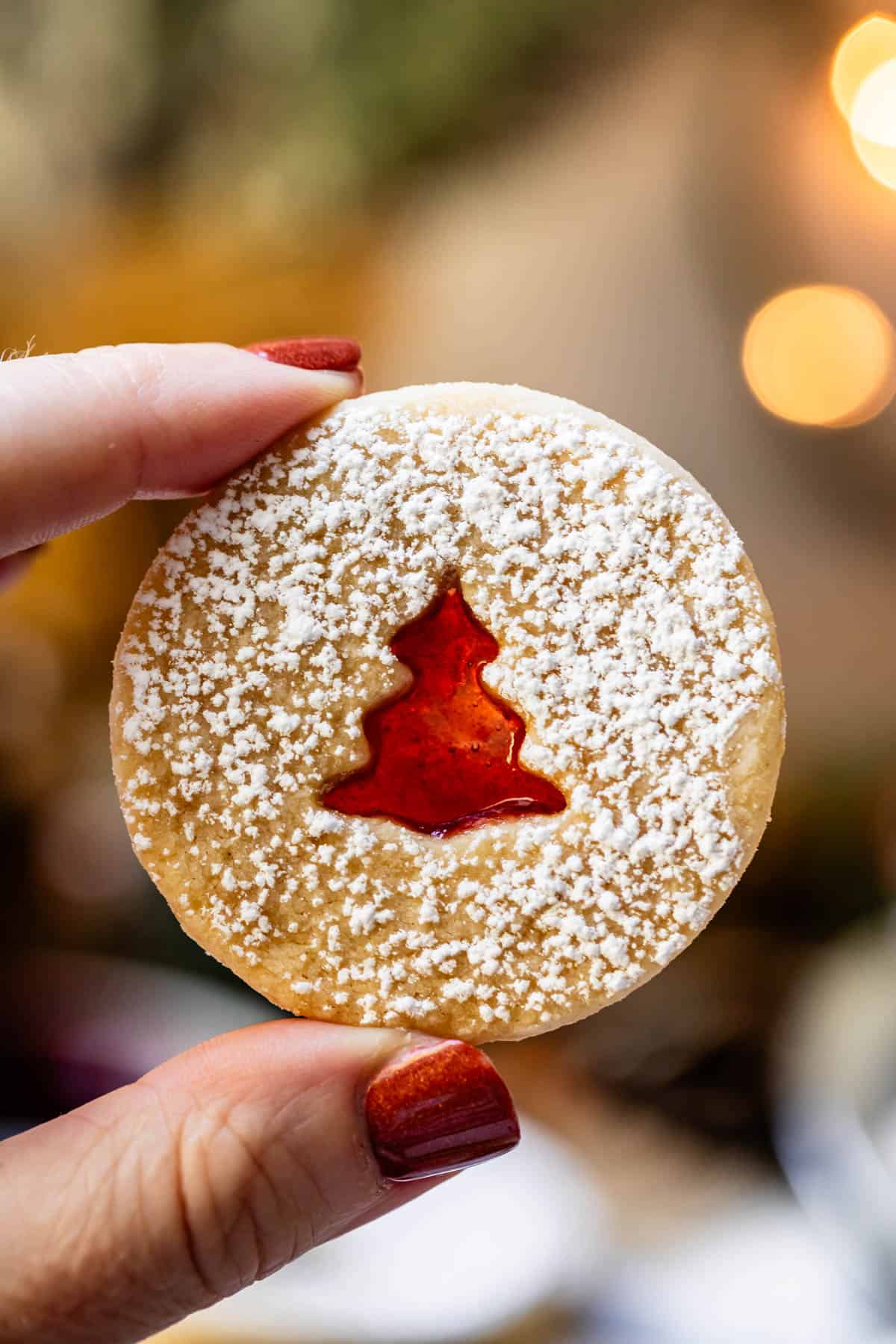 a finger and thumb holding a linzer cookie with a tree cutout showing jam.