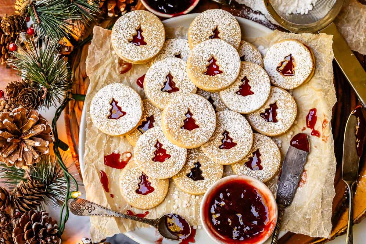 a platter stacked with nearly 20 linzer cookies, with a bowl of jam and pinecone garlnd nearby.