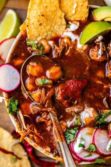 metal spoon in a bowl of pozole rojo with tostada, radish, and lime garnish.