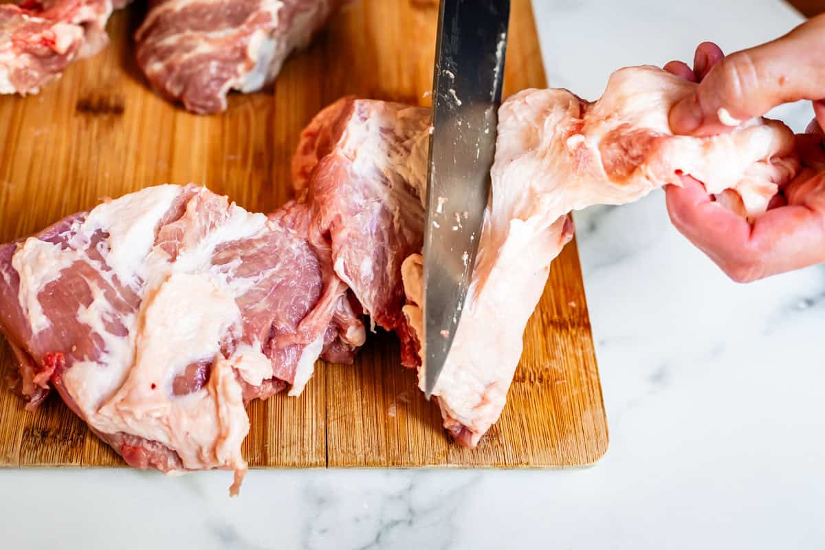 a chefs knife cutting a large piece of fat off pork shoulder on a wooden cutting board.