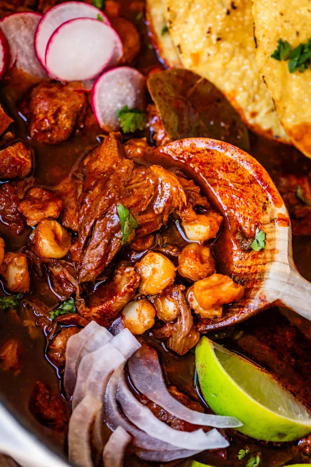 wooden spoon stirring pozole in a bowl with pork, hominy, onion, radish, and tostada.