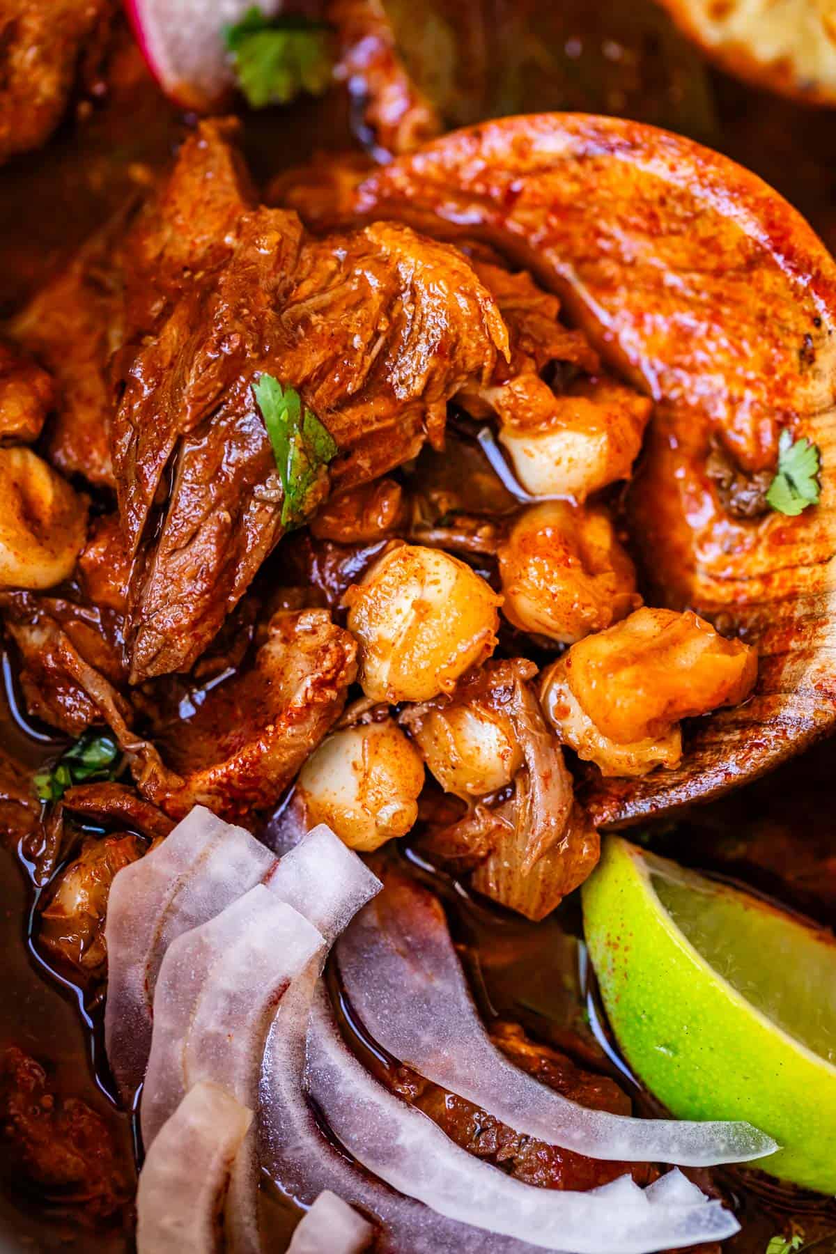wooden spoon lifting a bite of pork pozole with hominy and onion.
