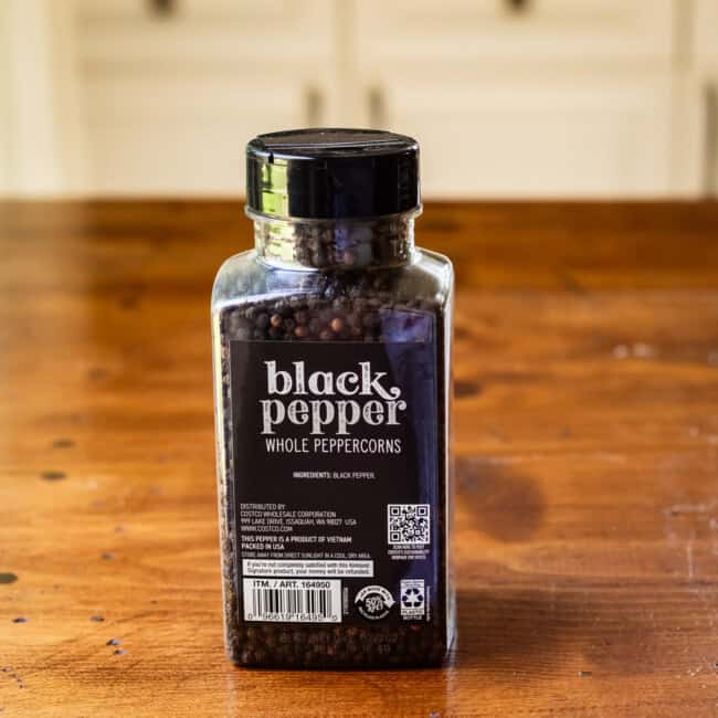 Container of whole black peppercorns on wooden table.