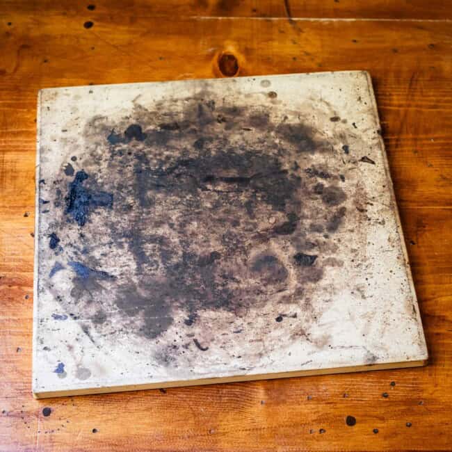 Well-used square pizza stone with charred stains on wooden table.