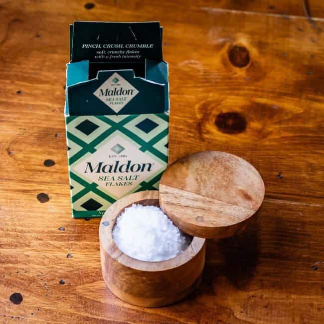 Open box of Maldon sea salt flakes with salt container on wooden table.