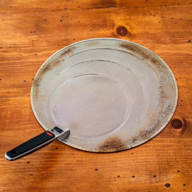 Stainless steel splatter shield with foldable handle on wooden table.