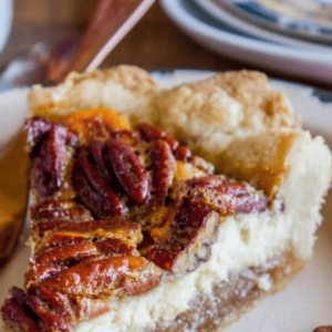 A close-up of a pecan pie cheesecake slice with a thick crust, creamy cheesecake layer, and caramelized pecans on top. A few pecan halves are scattered nearby on the plate.