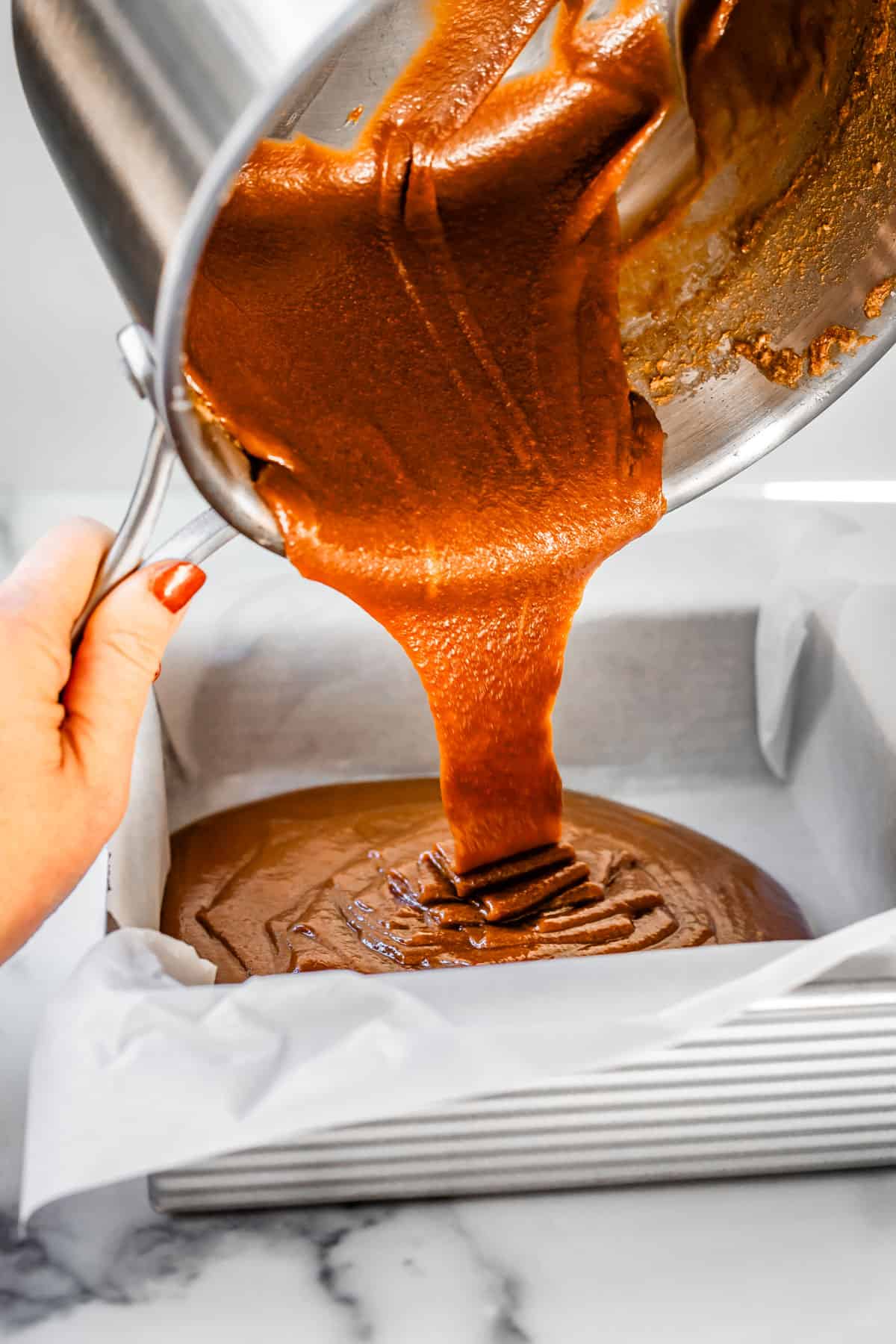 hand holding a metal pot, pouring hot caramel into a parchment lined square pan.