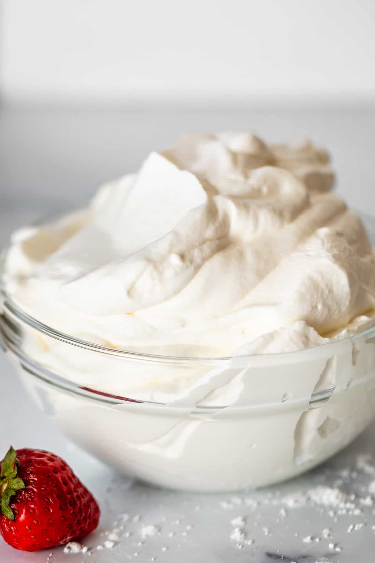 glass bowl of fluffy whipped cream on a counter with a single strawberry next to it.