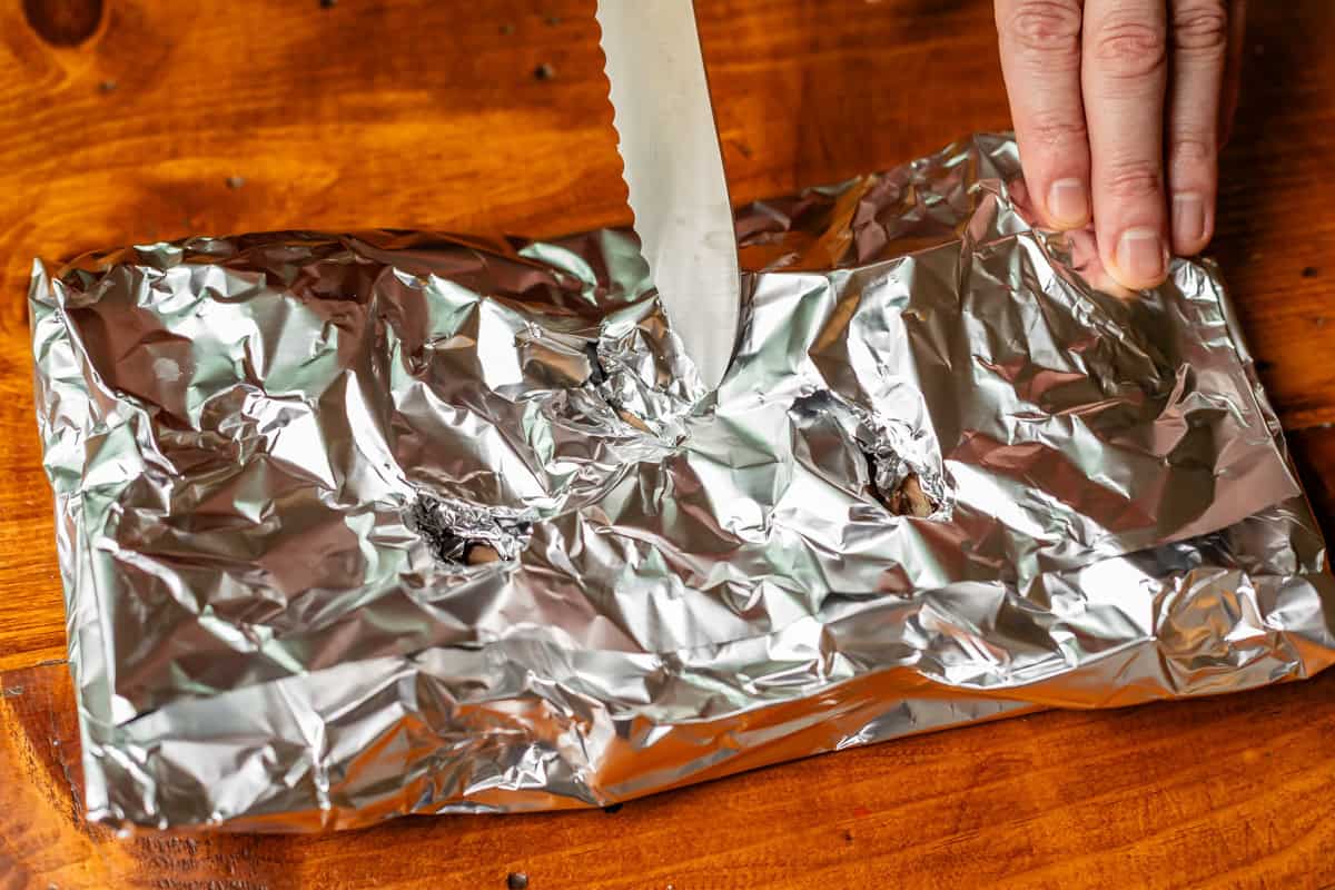 hand holding down a foil pouch filled with wood chips while knife cuts a slit in it.