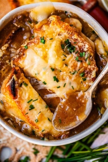 metal spoon holding french onion soup above bowl with toasty bread and cheese.