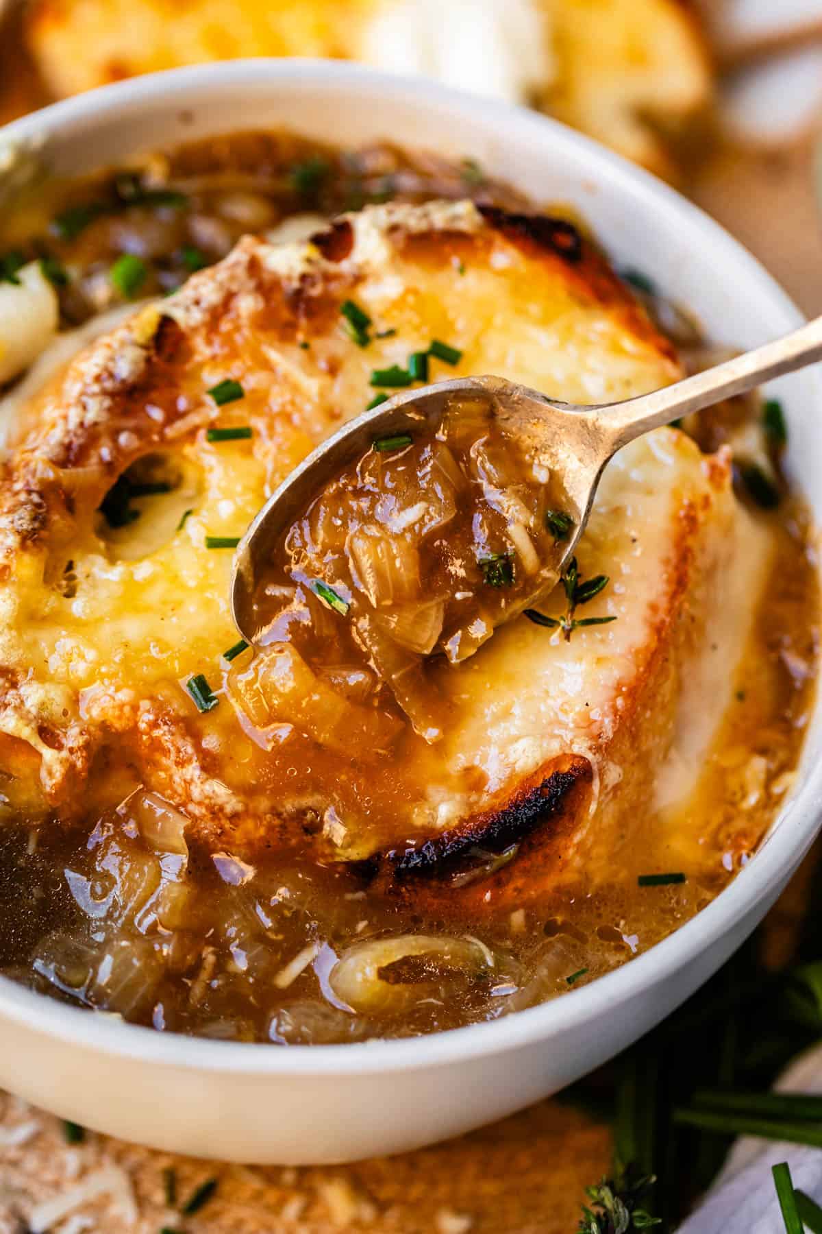 metal spoon holding french onion soup above bowl with toasty bread and cheese.