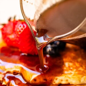 Homemade maple syrup being poured out of a glass spout over pancakes with strawberries in the background.