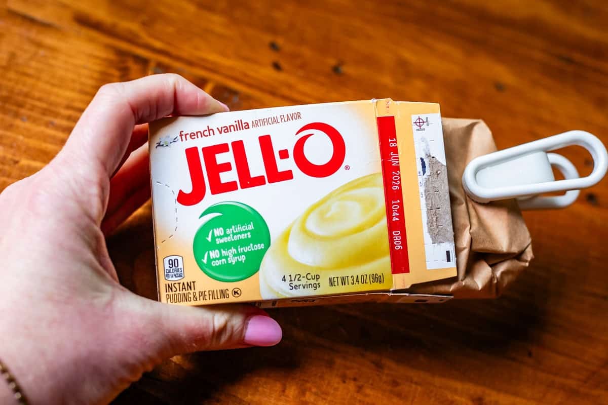hand holding a bow of instant vanilla pudding mix on a wood table.