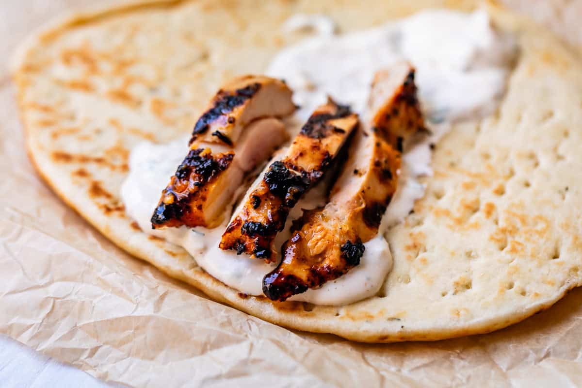 naan on parchment paper topped with garlic white sauce and three grilled chicken slices.