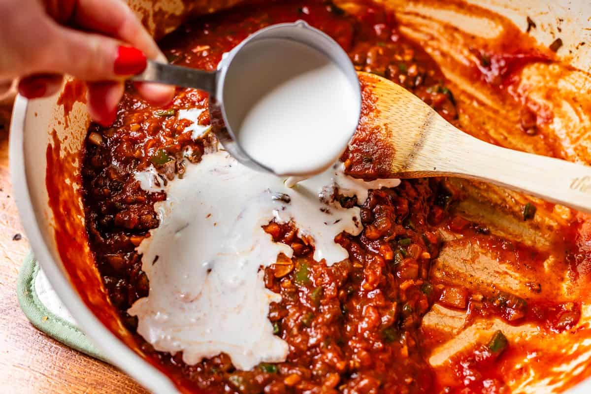 hand pouring cream from a measuring cup into a pan with sauce.