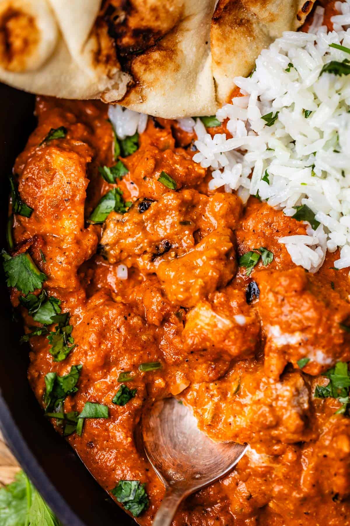 metal spoon digging into tikka masala with rice and naan next to it.