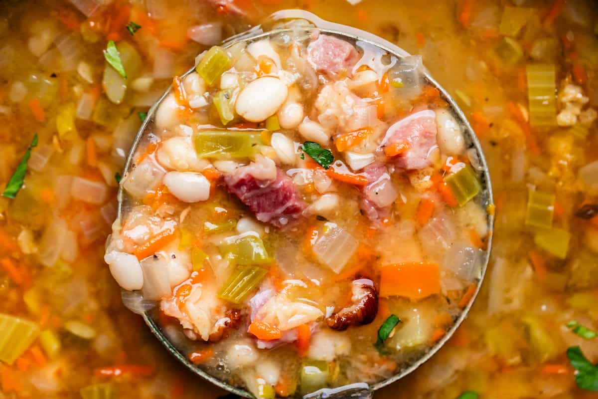 ladle scooping up a large amount of ham & bean soup showing the ham, beans, & veggies.