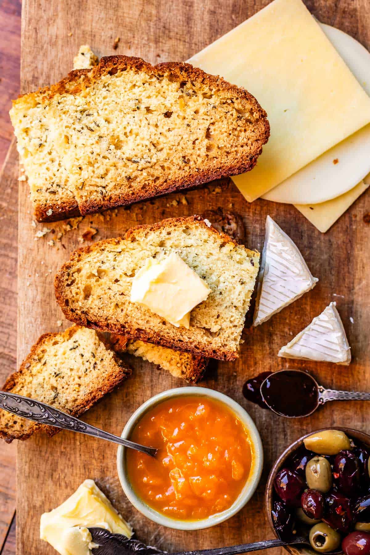 Delicious spread of bread board with Irish soda bread, apricot jam, cheeses, and olives on a wooden cutting board.