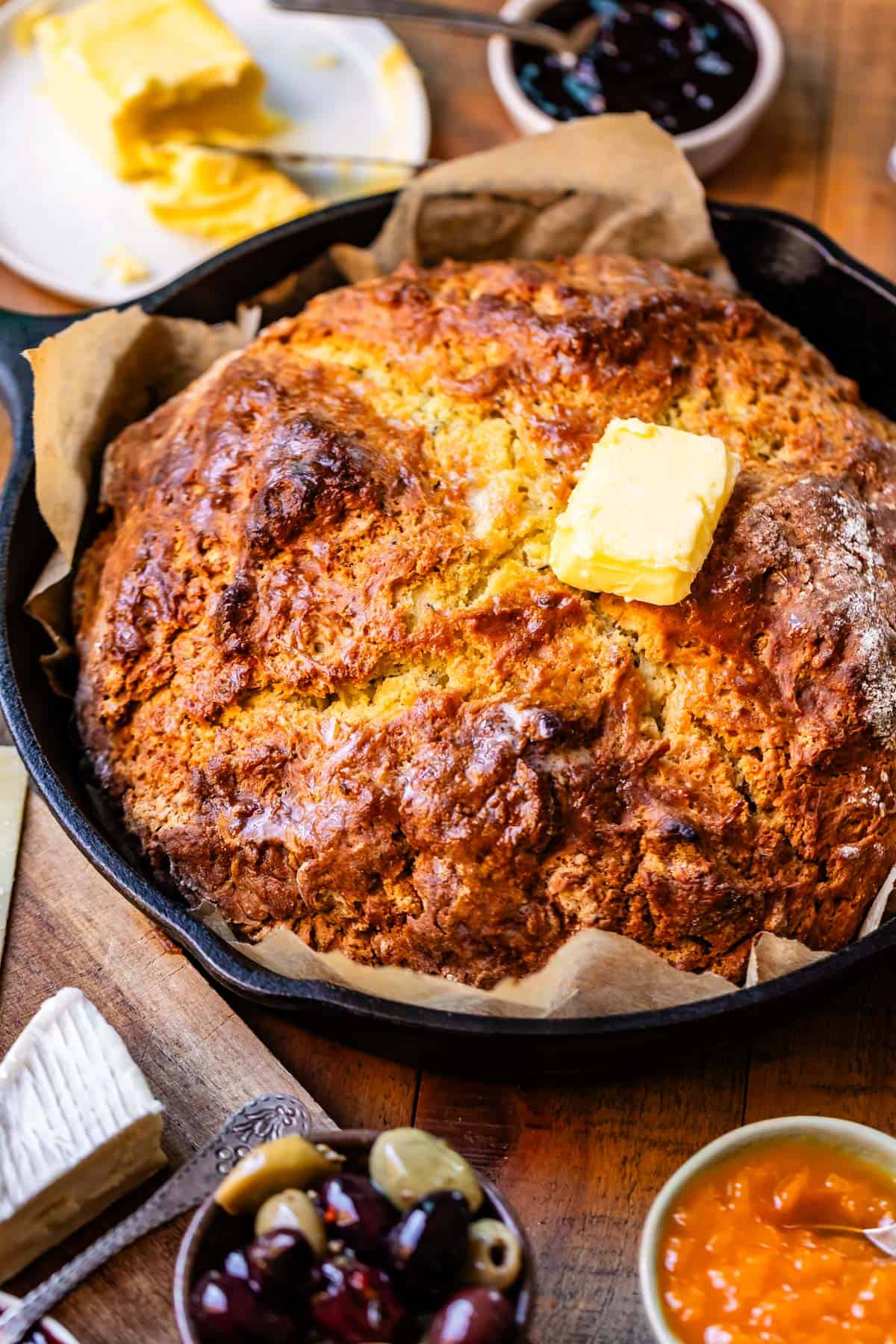 Baked round loaf of Irish soda bread with golden brown crusty top on parchment paper in cast iron skillet with toppings surrounding including butter, jam, and olives.