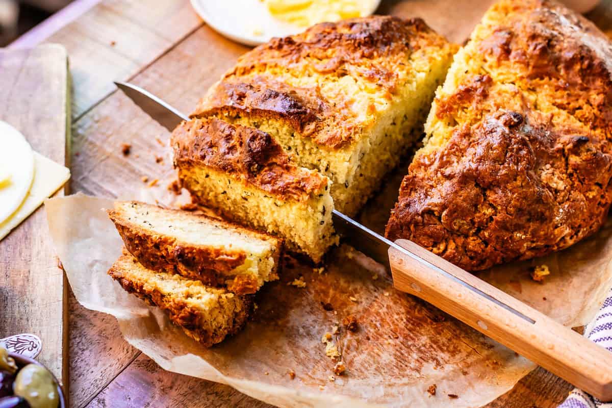 Round loaf of Irish soda bread in halves being sliced with bread knife on parchment paper on top of wooden cutting board.