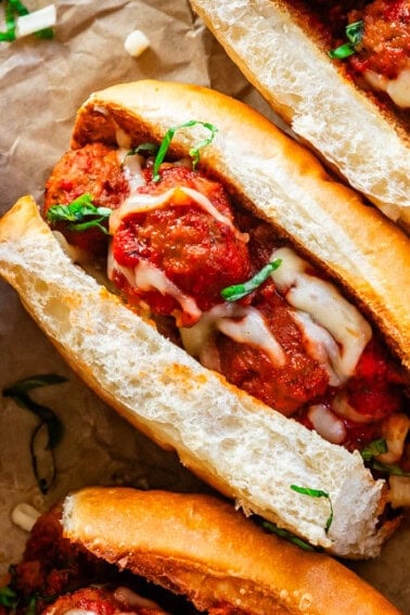 Several meatball subs laid out on parchment paper with cheese and parsley sprinkled on top.