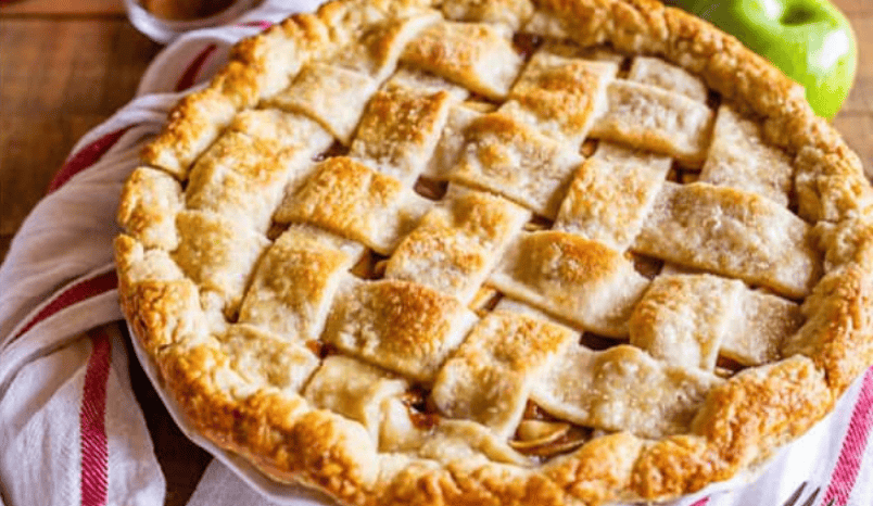 A freshly baked apple pie with a golden brown, lattice crust sits on a white dish, with a red-and-white striped towel and a green apple in the background.