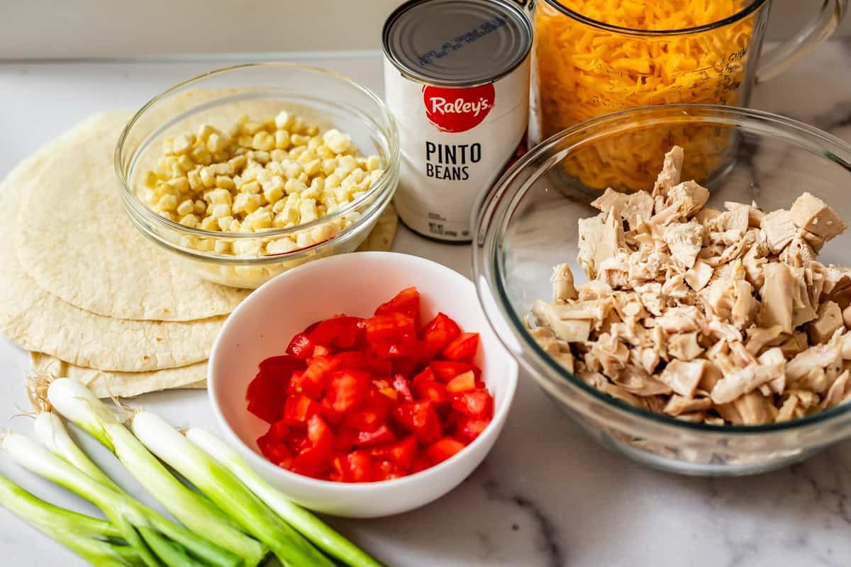 A marble countertop with tortillas, chopped green onions, diced tomatoes, cooked corn, shredded cheddar cheese, a can of pinto beans, and a bowl of chopped cooked chicken.