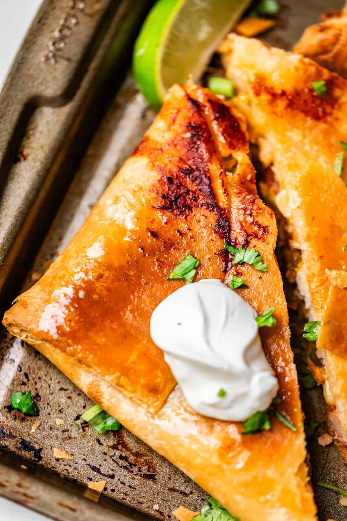 A close-up of a golden, crispy quesadilla slice topped with a dollop of sour cream and chopped cilantro, served on a baking tray with a lime wedge in the background.