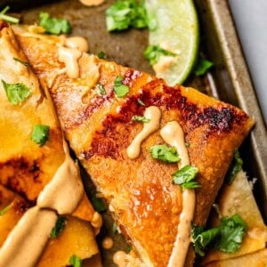 A close-up of crispy, golden quesadillas drizzled with a creamy sauce, garnished with chopped cilantro, and served with a wedge of lime on a baking tray.