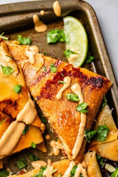 A close-up of crispy, golden quesadillas drizzled with a creamy sauce, garnished with chopped cilantro, and served with a wedge of lime on a baking tray.