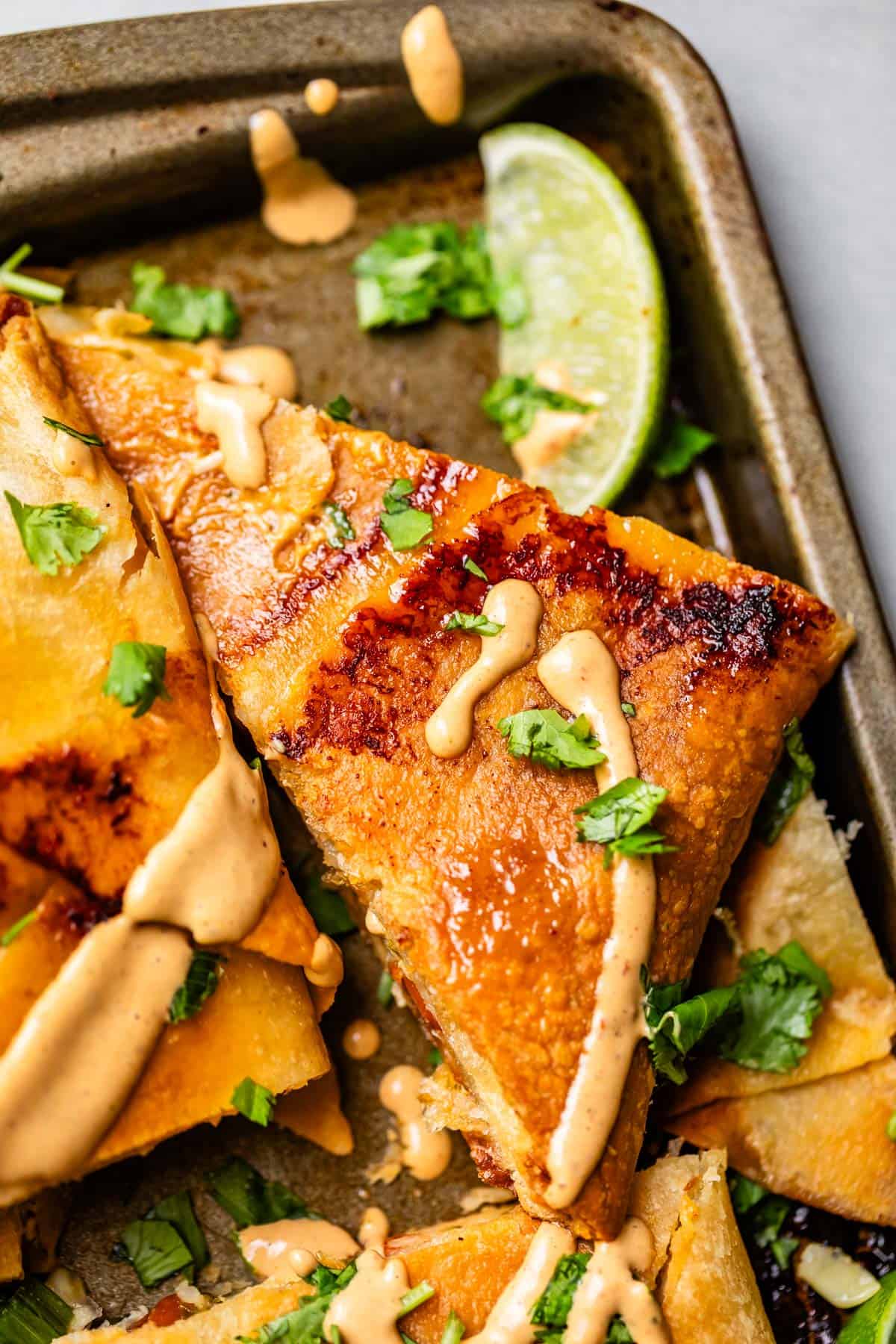 A close-up of crispy, golden quesadillas drizzled with a creamy sauce, garnished with chopped cilantro, and served with a wedge of lime on a baking tray.
