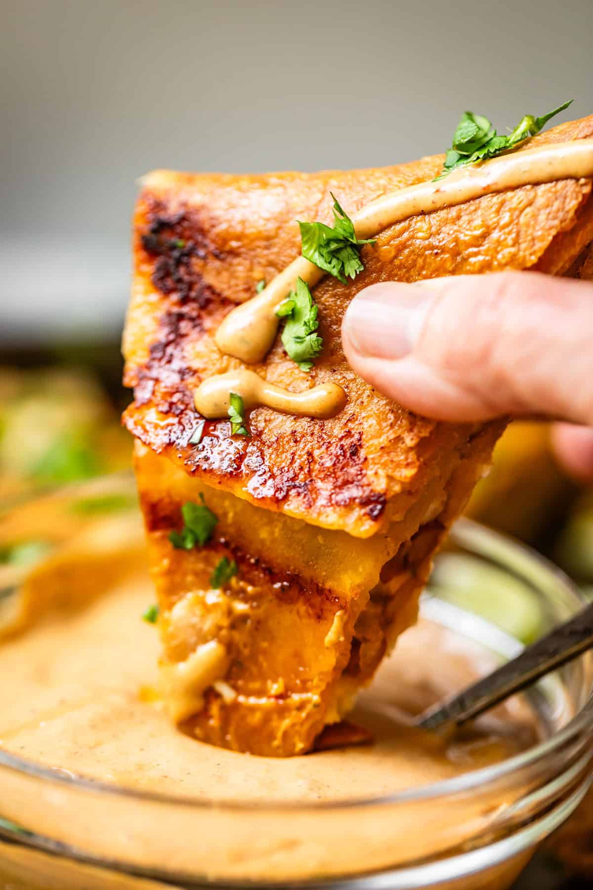 A hand holds a slice of cheesy quesadilla topped with herbs and a sauce drizzle, dipping it into a bowl of creamy dipping sauce.