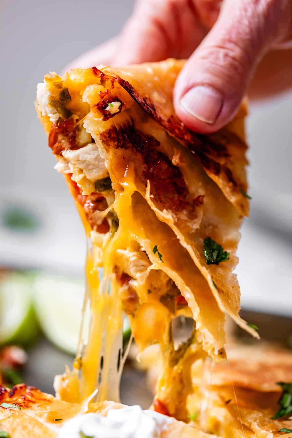A close-up of a hand pulling a cheesy quesadilla slice, with melted cheese stretching and bits of vegetables and herbs visible inside.