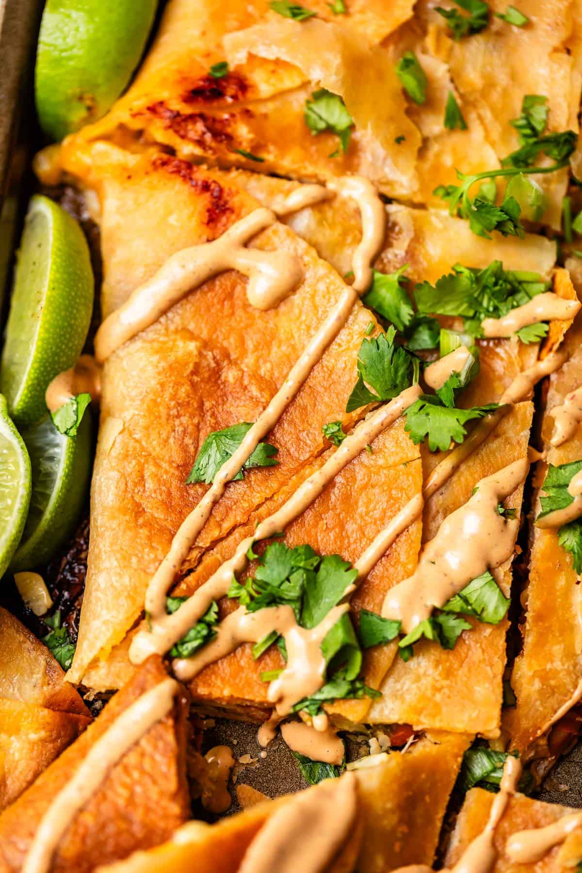 Close-up of golden, crispy quesadillas cut into triangles, drizzled with creamy sauce, garnished with fresh cilantro, and served with lime wedges.
