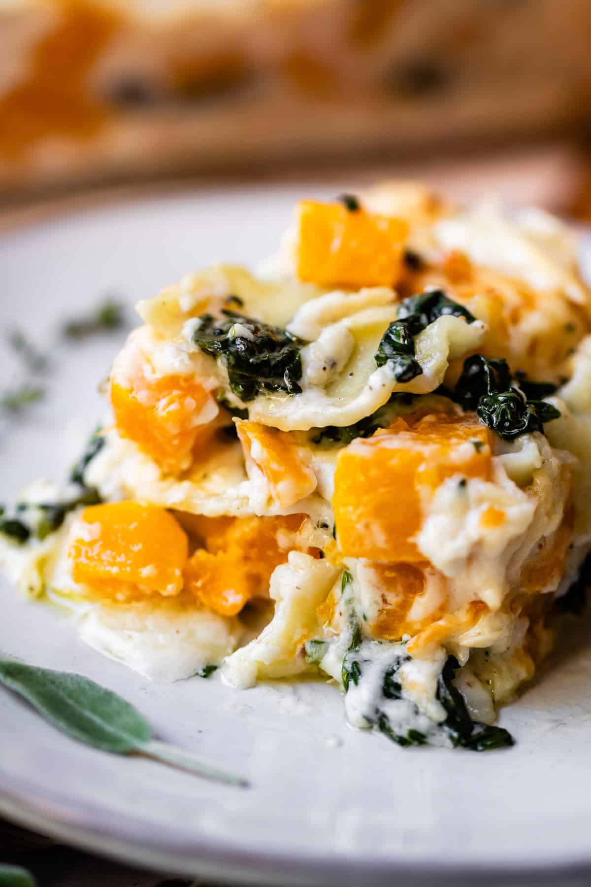 A close-up of a serving of creamy butternut squash and spinach lasagna on a white plate, showing layers of pasta, orange squash, wilted spinach, and melted cheese.