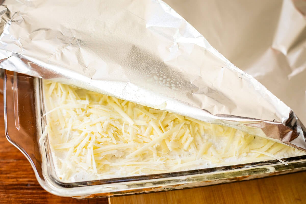A glass baking dish filled with shredded cheese and a creamy mixture, partially covered with a sheet of aluminum foil, sits on a wooden surface.