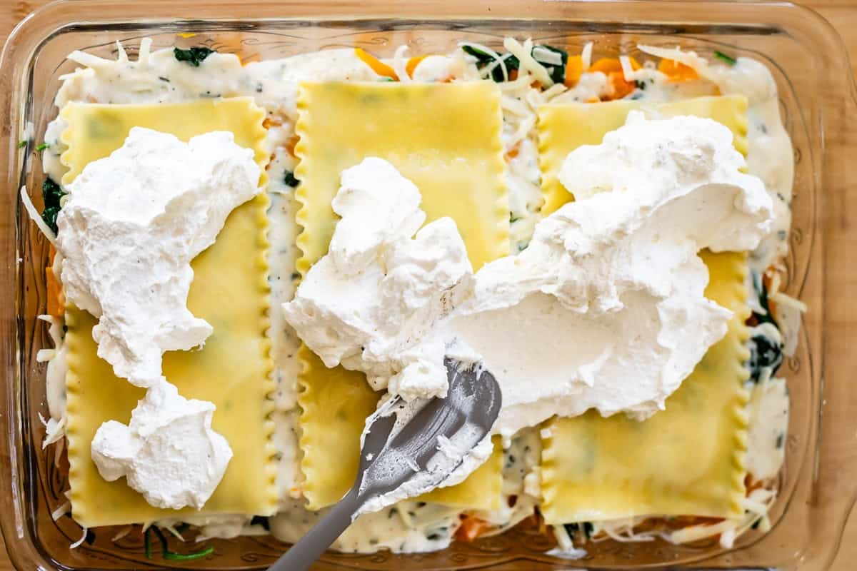 A close-up of a baking dish filled with uncooked lasagna. A spatula spreads a thick layer of ricotta cheese over pasta sheets, with visible spinach, sauce, and shredded cheese underneath.