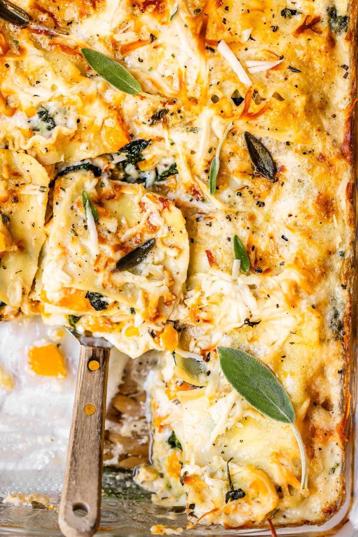 A close-up of cheesy baked ravioli with spinach and herbs in a casserole dish, with a serving spatula scooping out a portion. Fresh sage leaves are visible on top.