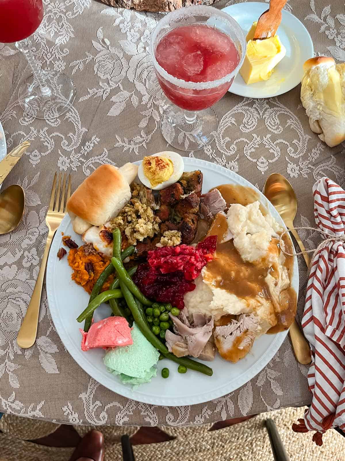 A plate filled with Thanksgiving food including turkey with gravy, mashed potatoes, stuffing, cranberry sauce, green beans, peas, sweet potatoes, a dinner roll, a deviled egg, and colorful desserts, with a pink drink beside it.