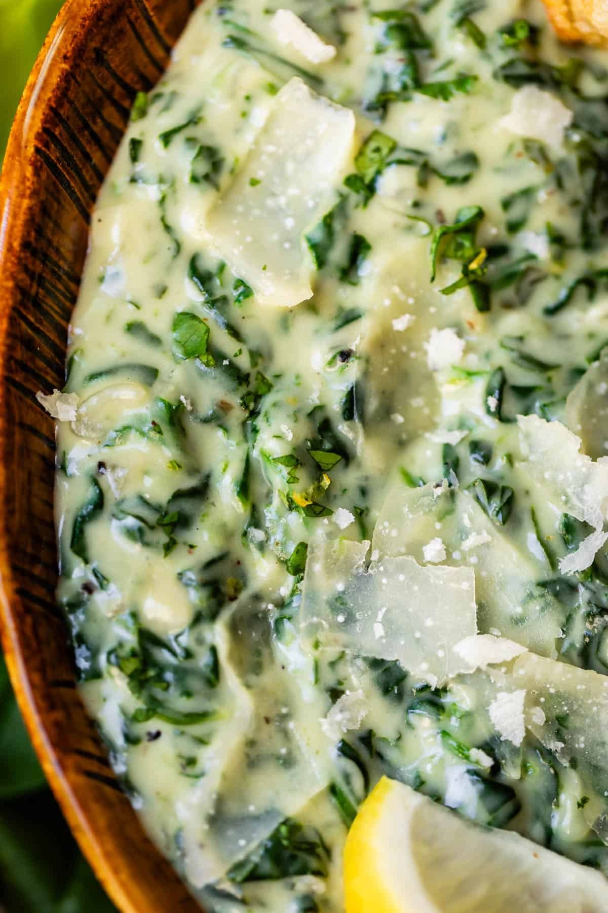 Close-up of creamy spinach dip in a wooden bowl, topped with shaved parmesan cheese and a lemon wedge. The dip has a rich, smooth texture with visible bits of spinach.