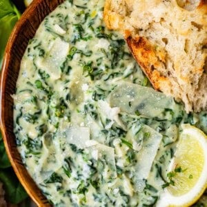 A wooden bowl filled with creamy spinach dip topped with Parmesan shavings, served with a slice of rustic bread and a wedge of lemon on the side.
