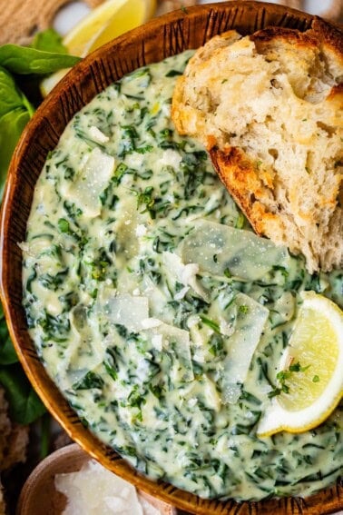 A wooden bowl filled with creamy spinach dip topped with Parmesan shavings, served with a slice of rustic bread and a wedge of lemon on the side.