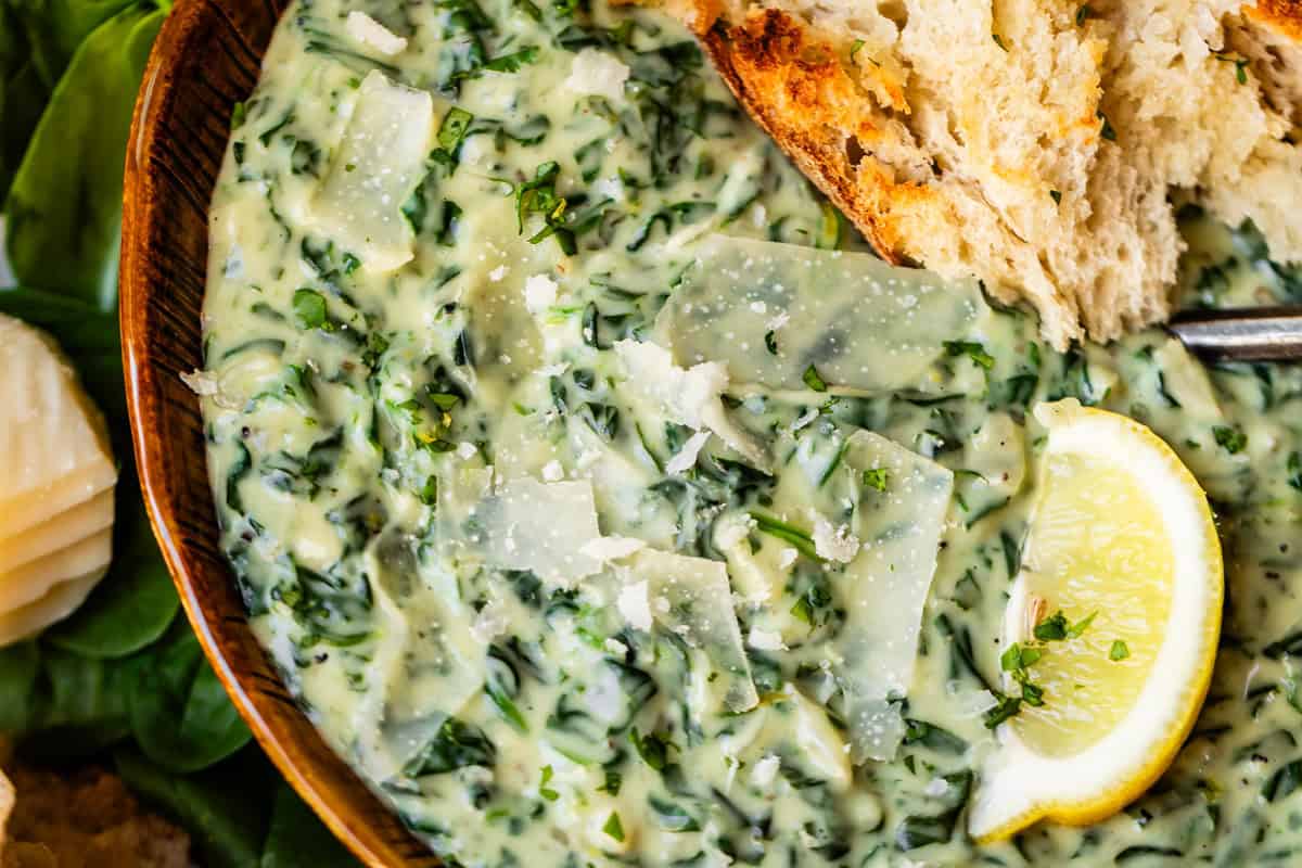 A close-up of creamy spinach artichoke dip topped with shaved parmesan, served with a slice of crusty bread and a lemon wedge.