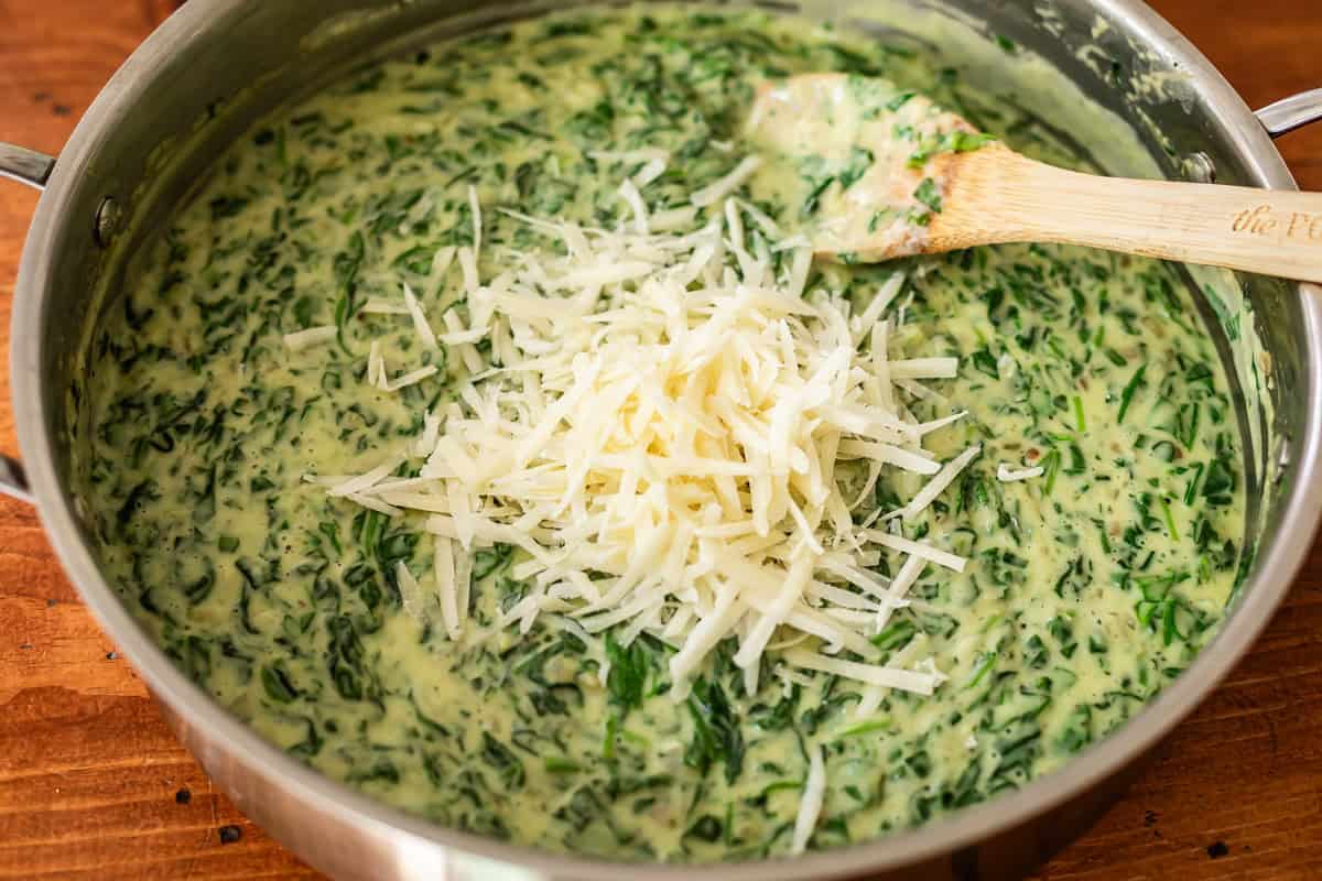 A pan filled with creamy spinach dip is topped with a pile of shredded cheese, and a wooden spoon rests inside the mixture. The pan sits on a wooden surface.