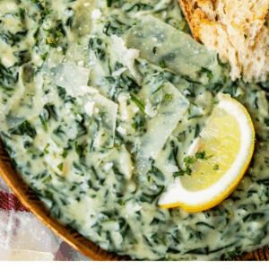 A wooden bowl filled with creamy creamed spinach, topped with shaved parmesan, a lemon wedge, and served with a piece of crusty bread. Text at the bottom reads: Rich & Creamy Creamed Spinach - The Food Charlatan.