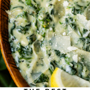 Close-up of creamy, cheesy spinach in a wooden bowl, garnished with shaved parmesan and a lemon wedge. Text at the bottom reads: The Best Creamed Spinach of Your Life, The Food Charlatan.