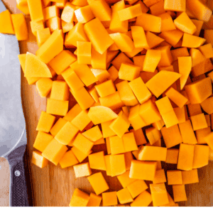 Diced butternut squash pieces on a wooden cutting board next to a knife, with text that reads, The very best way to cut butternut squash. The Food Charlatan.
