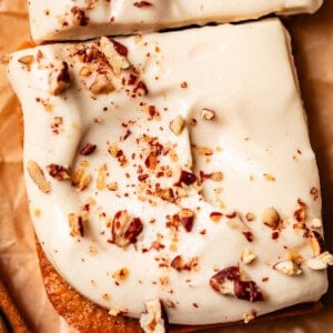 HERO? Close-up of a frosted dessert bar topped with chopped nuts, placed on brown parchment paper. Another similar bar is partially visible above, and a cinnamon stick rests nearby.